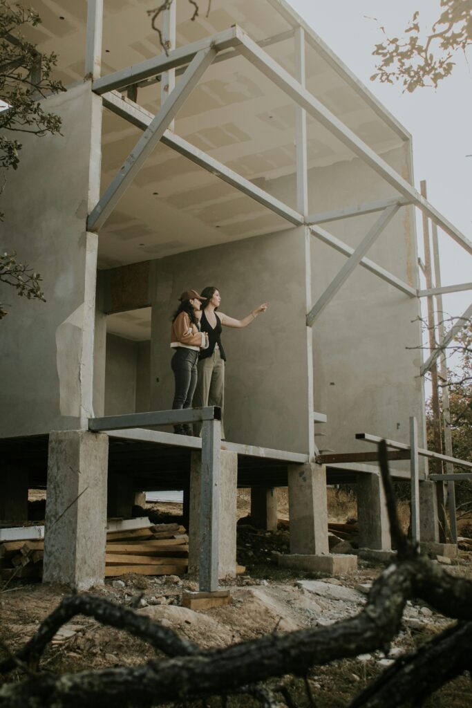 Two adults observe a modern concrete house under construction, surrounded by nature.